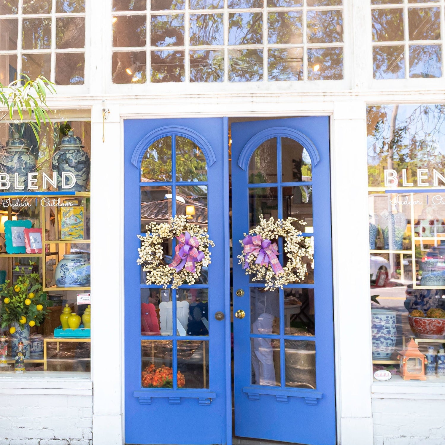 Blue double doors of a store named 'BLEND' with decorative wreaths, surrounded by plants and flowers.